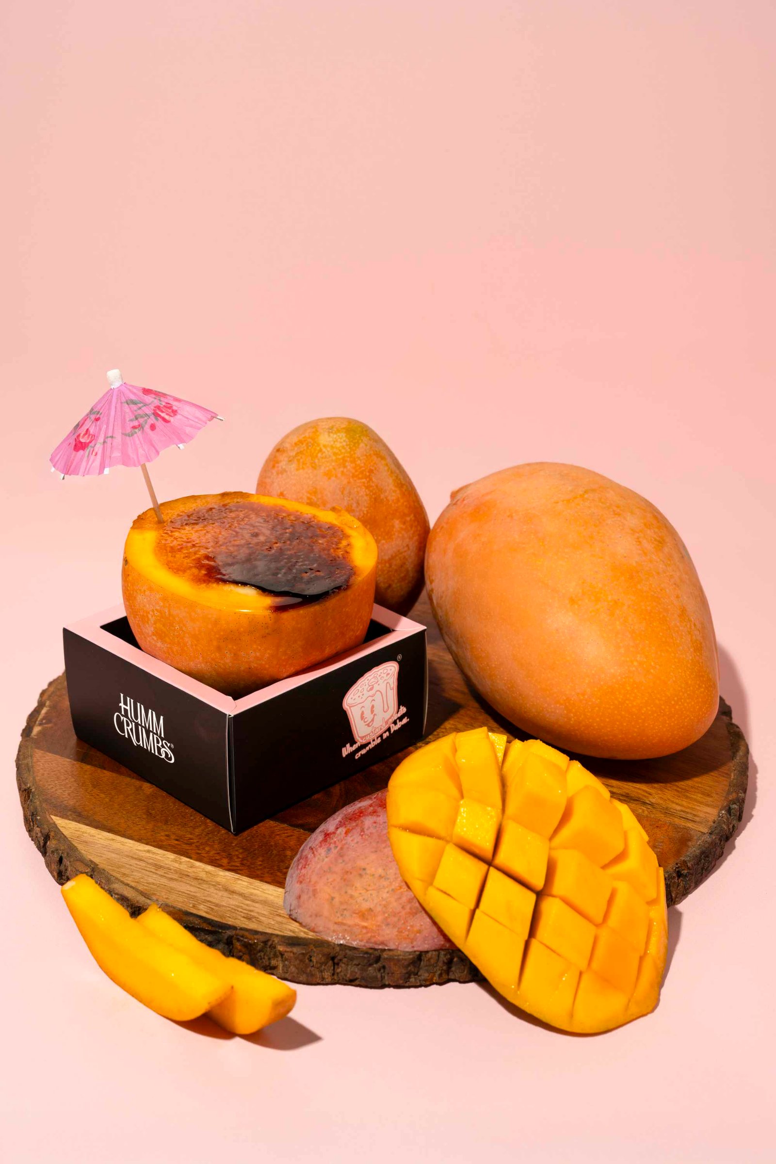 A mango half filled with torched custard, sitting in a Humm Crumbs box on a wooden board, surrounded by whole and sliced mangoes on a blush-pink ground.