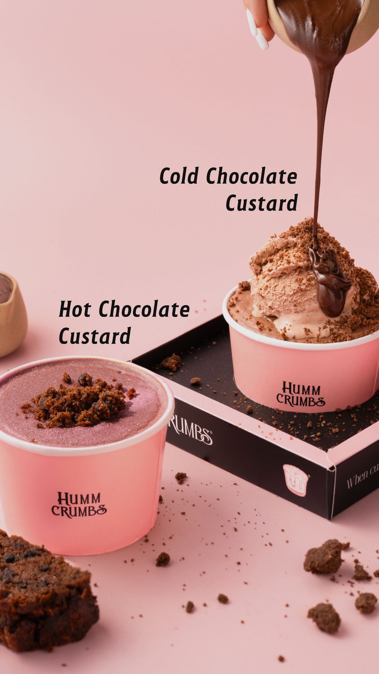 Two pink Humm Crumbs cups of chocolate custard, one hot and one cold, with melted chocolate being poured over the cold one and crumble scattered around.