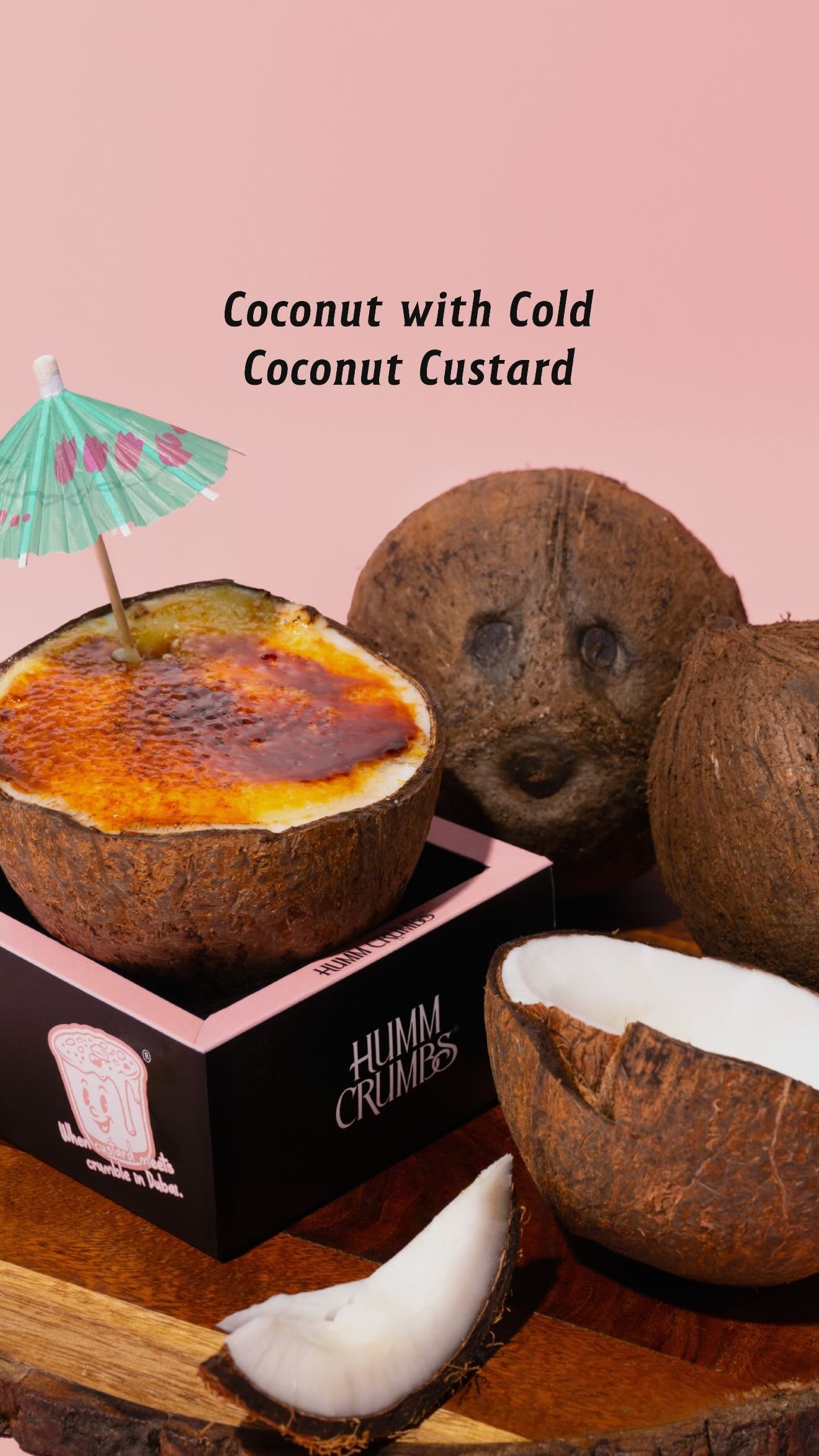 A coconut shell filled with torched coconut custard, sitting in a Humm Crumbs box beside whole and split coconuts.