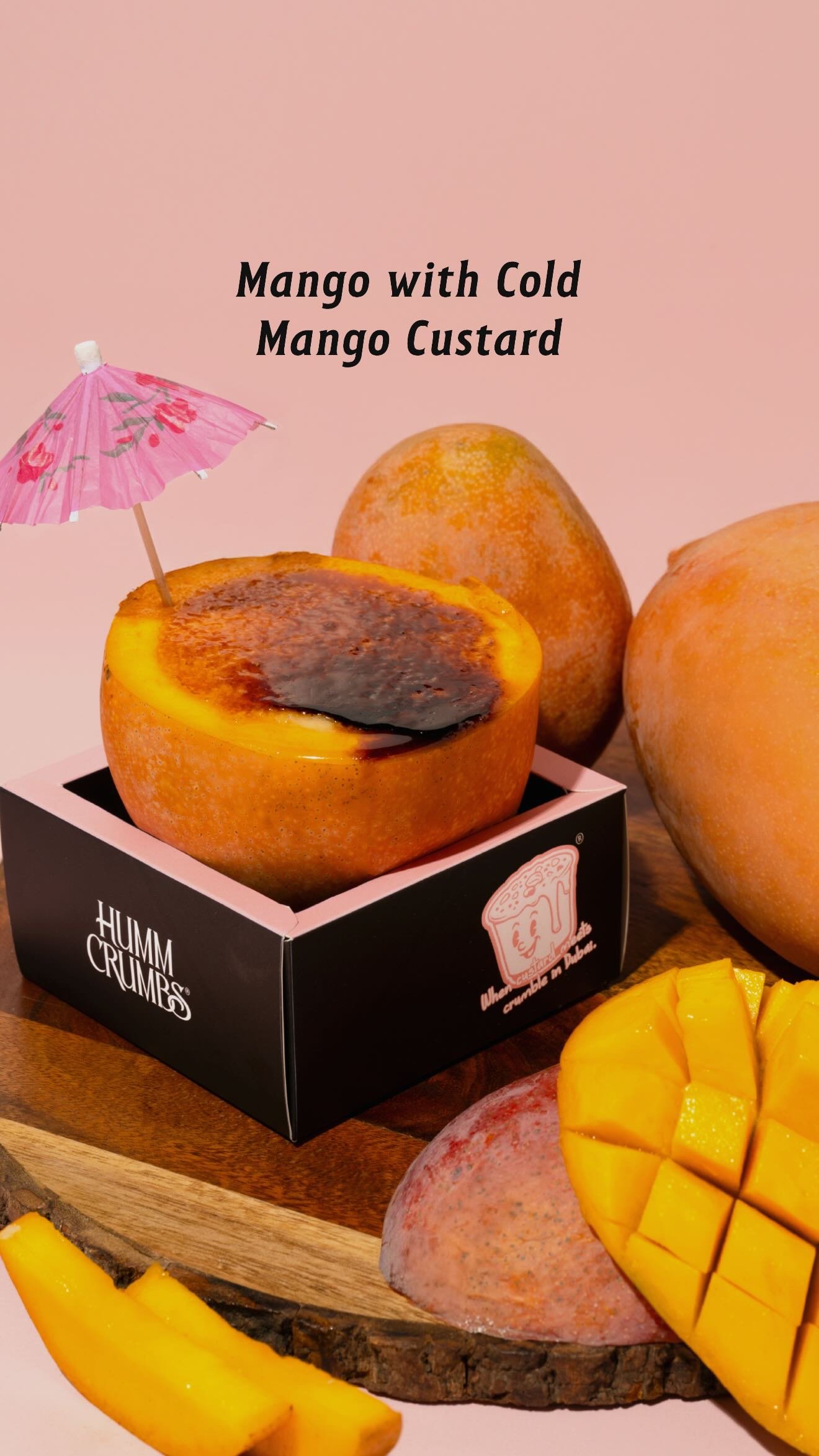 A mango half filled with torched mango custard, served in a Humm Crumbs box with whole and cubed mangoes alongside.