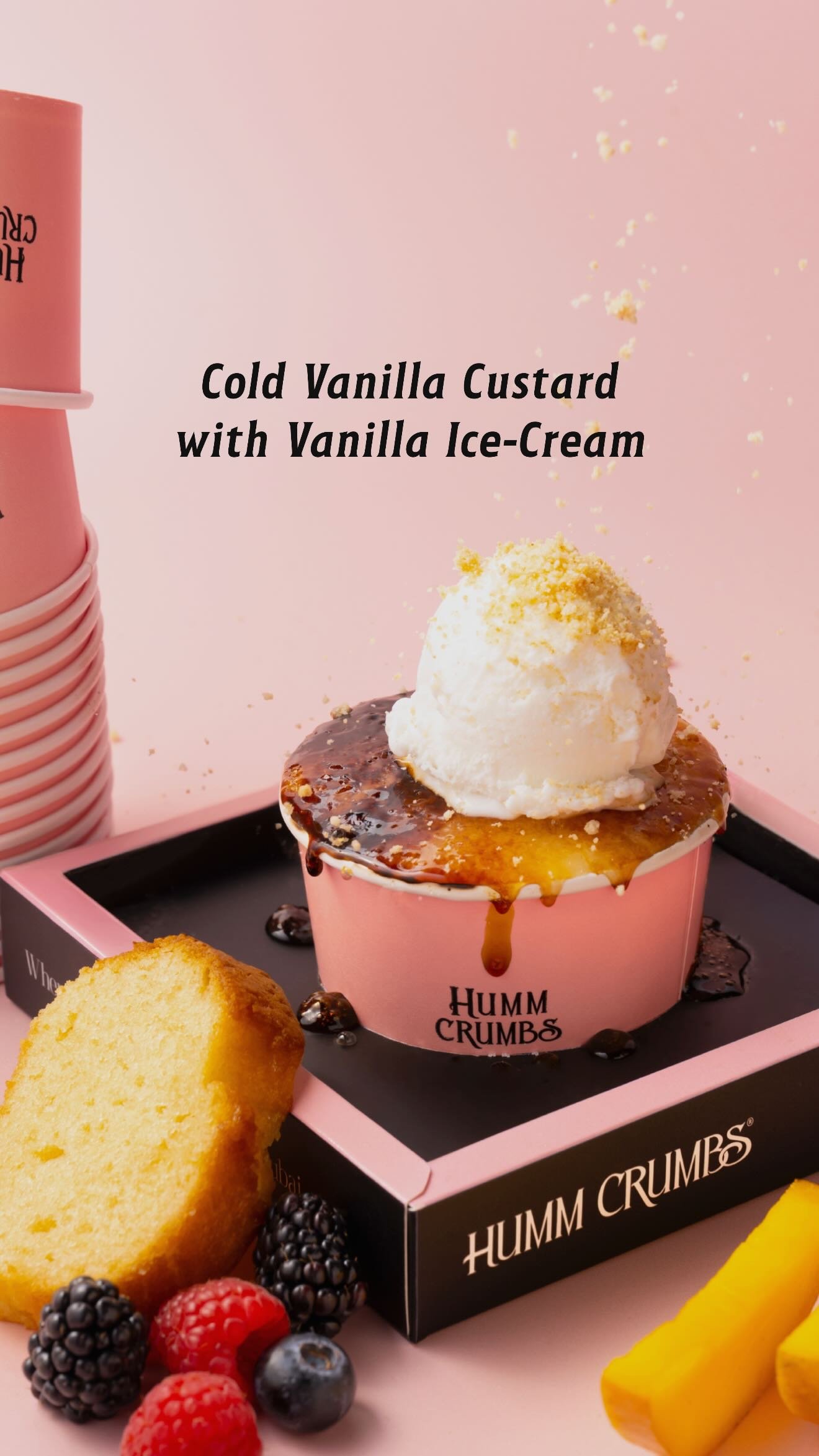 A pink Humm Crumbs cup of torched vanilla custard topped with a scoop of vanilla ice cream and crumble, beside a slice of milk cake and fresh berries.