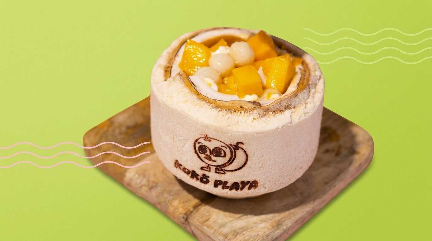 A halved coconut branded with the Koko Playa mascot, filled with panna cotta, mango cubes and mochi balls, on a wooden board against a lime-green ground.