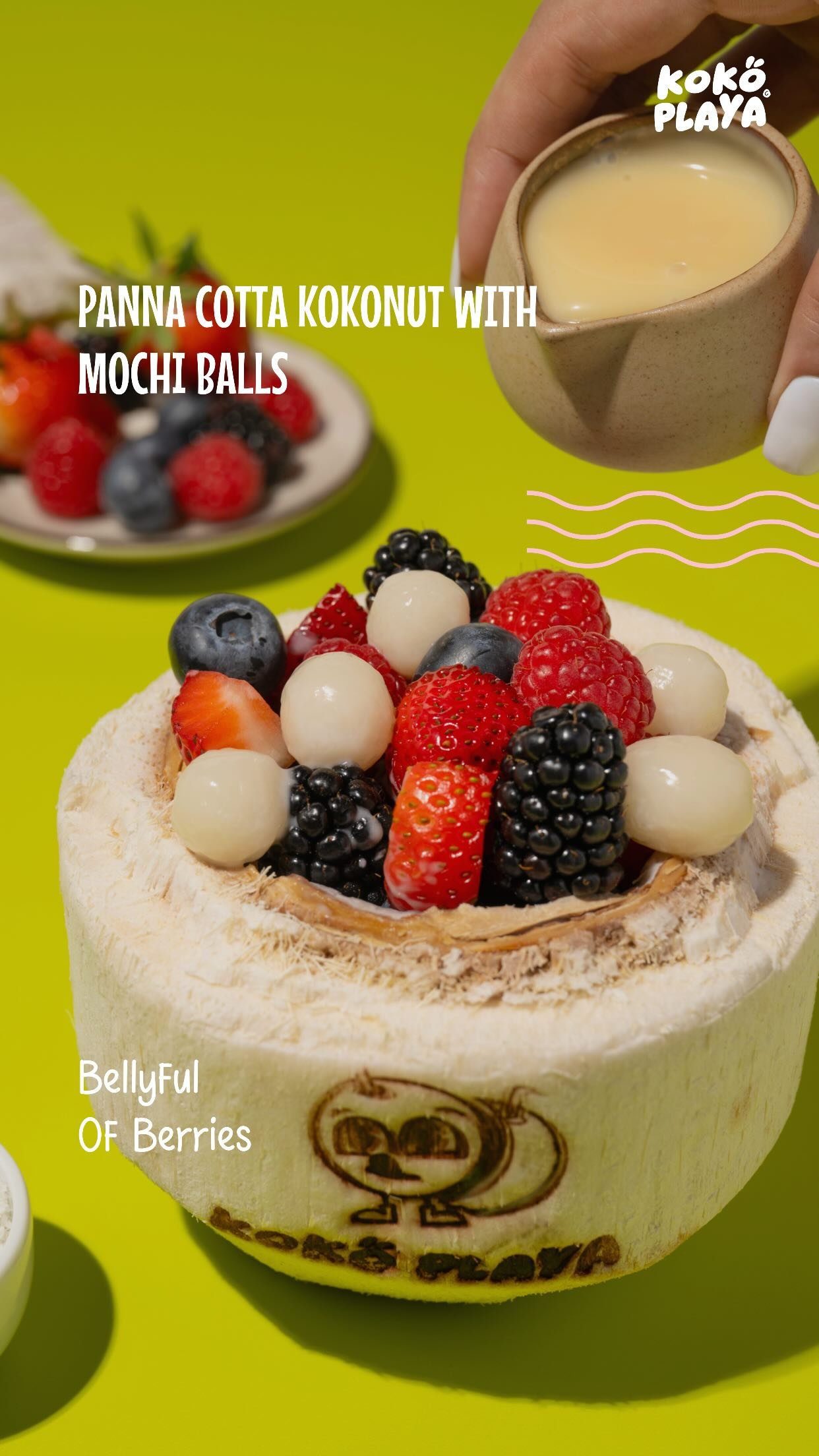 A branded coconut shell filled with panna cotta, mochi balls, strawberries, raspberries, blackberries and blueberries, with condensed milk being poured over.