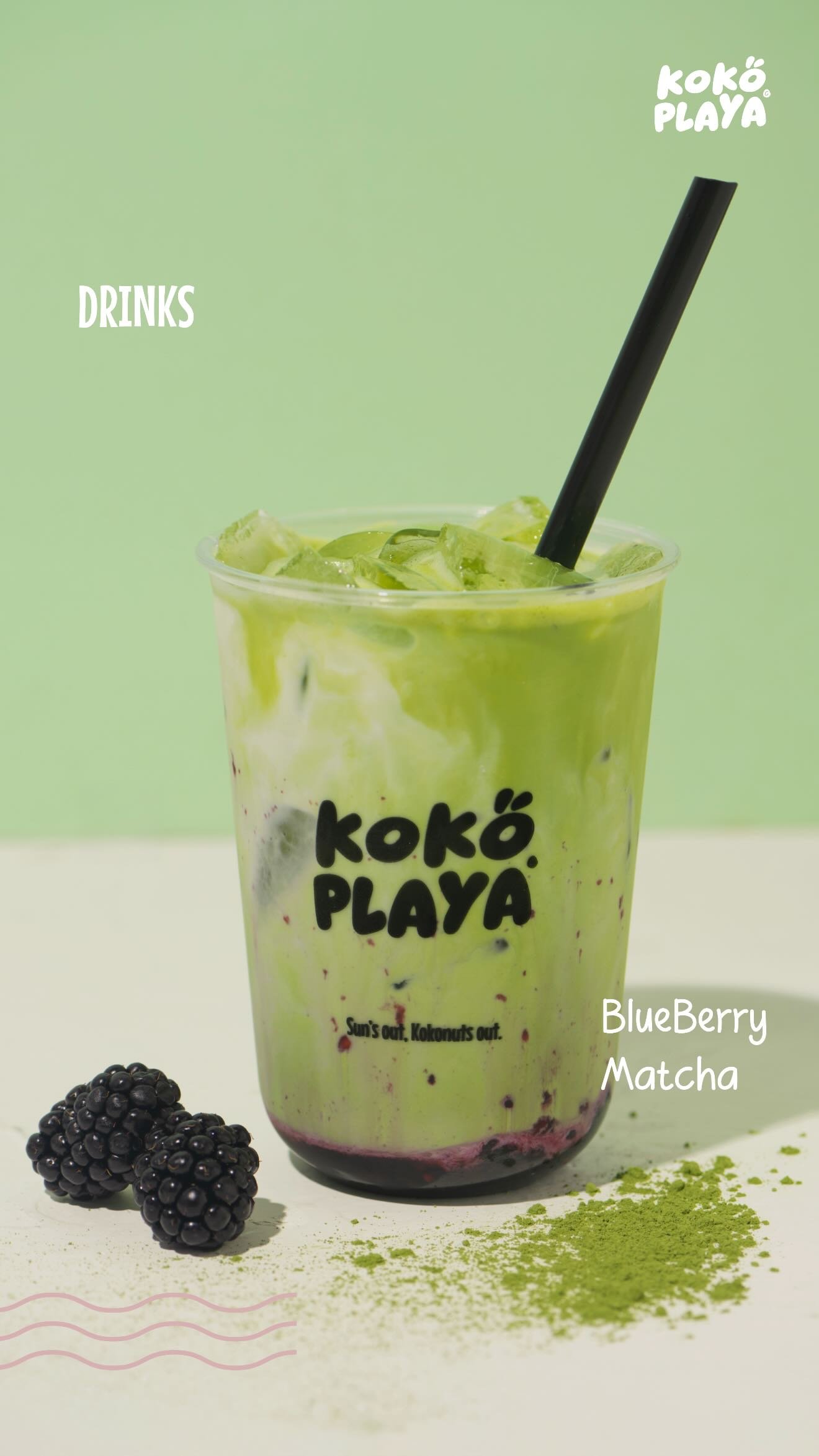 A clear Koko Playa cup of iced matcha layered over blueberry compote, beside fresh blackberries and a scatter of matcha powder.