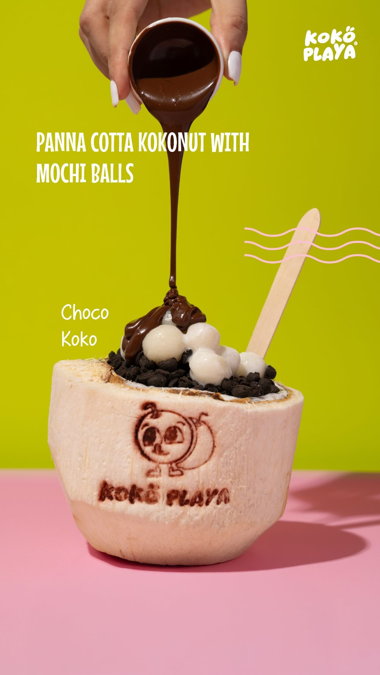 A branded coconut shell of panna cotta topped with mochi balls and dark cookie crumb, melted chocolate being poured over it from a small cup.