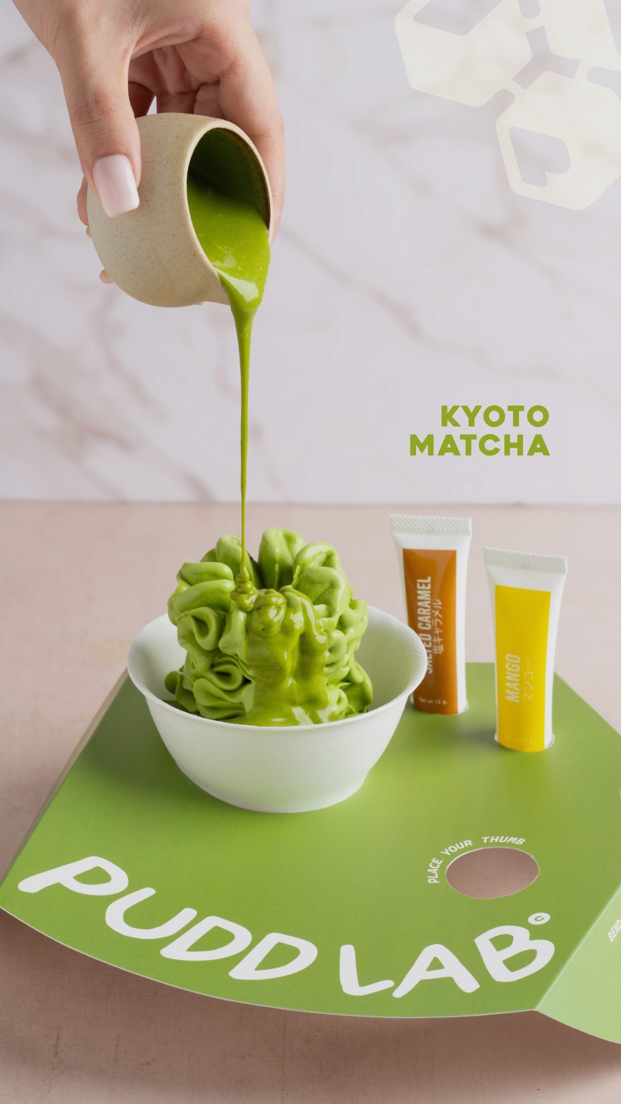 A white bowl of matcha soft serve on a green Pudd Lab tray, with matcha sauce being poured over it and branded sauce tubes standing alongside.